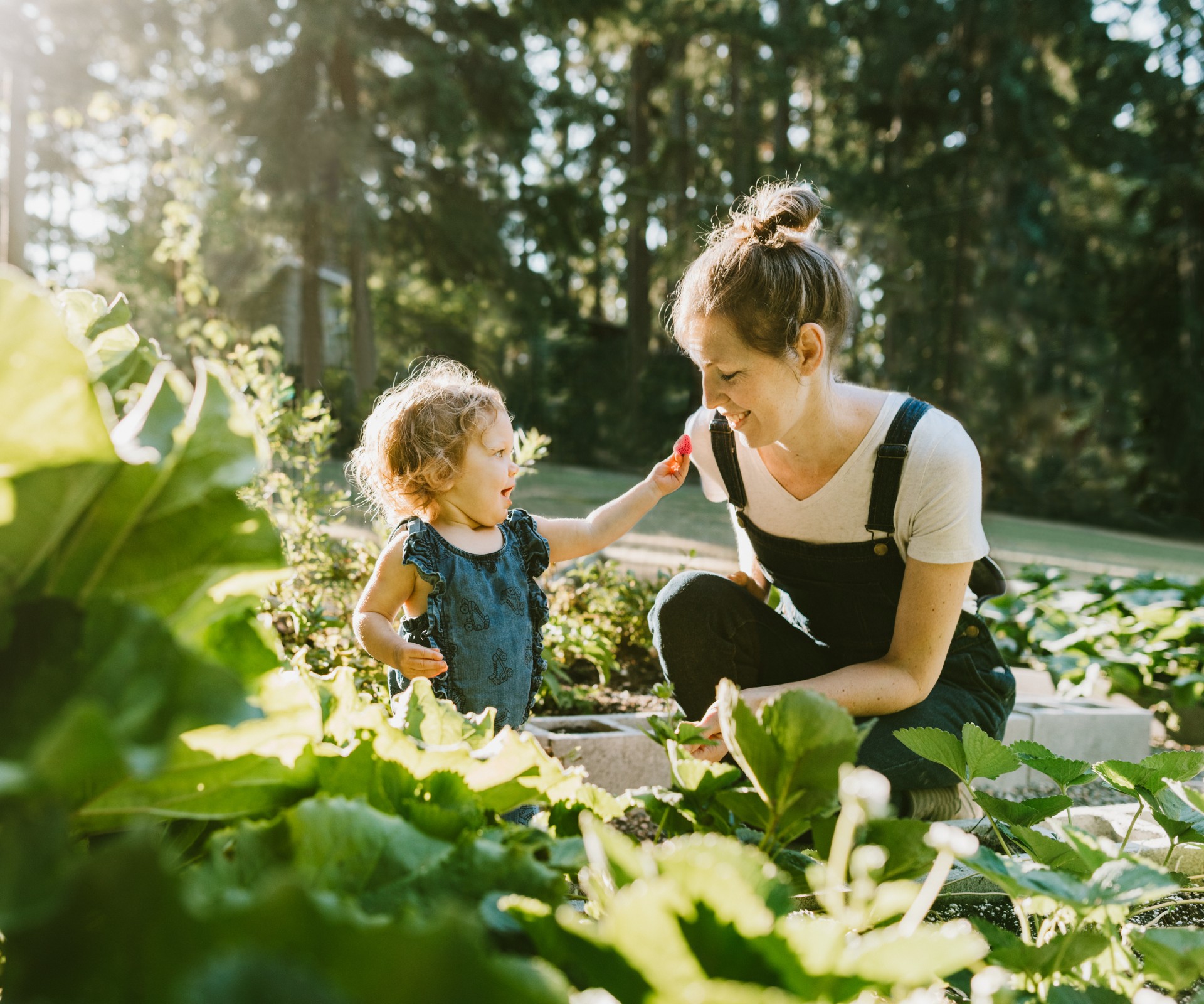 Lächelnde Mutter in einem grünen Beet mit ihrem kleinen Kind das ihr lächelnd eine Erdbeere entgegen streckt