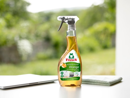 A Frosch multi-surface cleaner orange trigger bottle stands next to a cleaning cloth and laptop on a table