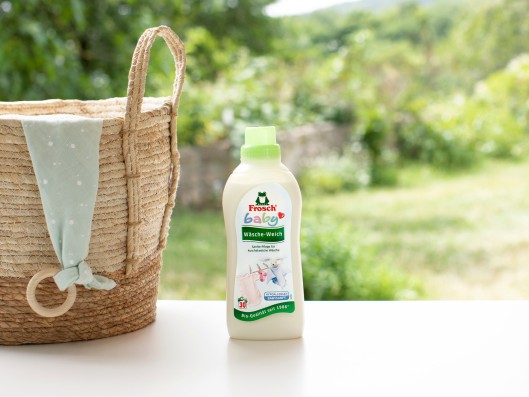 A bottle of Frosch Baby Fabric Softener stands next to a basket with cloth in front of a window