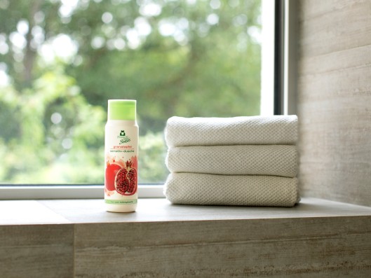In the bathroom in front of a window a Frosch Senses Pomegranate Sensitive Shower Gel bottle stands next to stacked towels